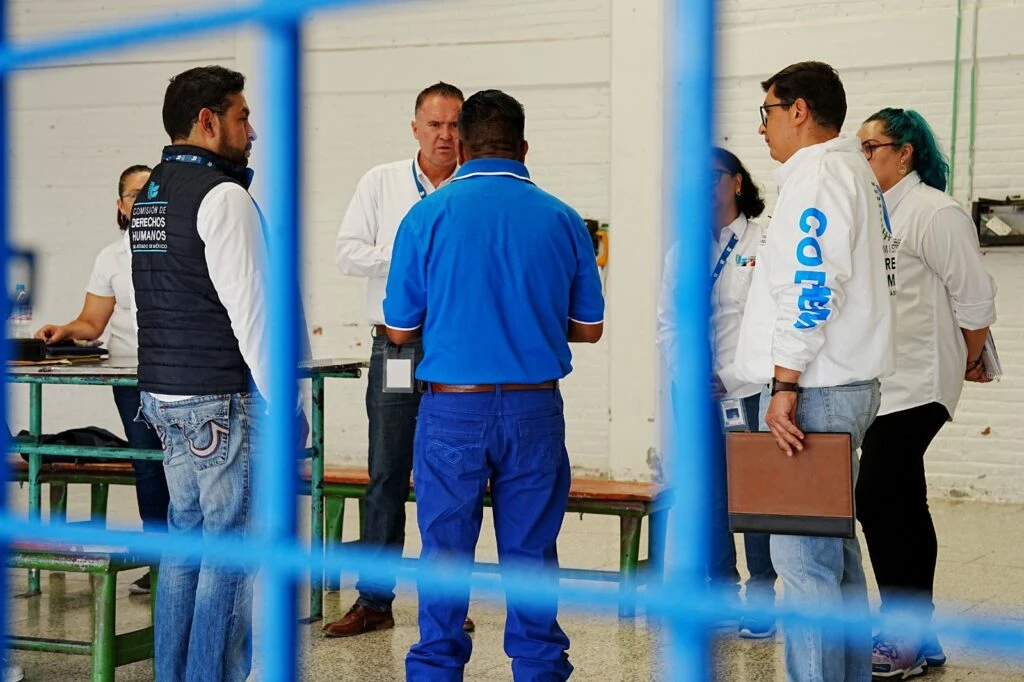 Varias personas de pie en un área interior tipo taller o instalación, conversando en grupo mientras portan vestimenta institucional y gafetes; al frente se aprecian estructuras metálicas desenfocadas.