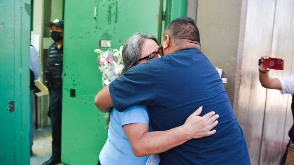 Dos personas se abrazan frente a una puerta metálica verde abierta, mientras otras personas observan alrededor; en el interior se aprecia presencia de personal de seguridad en un entorno institucional.