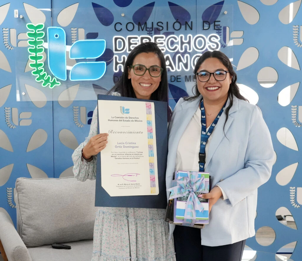 Persona expone frente a una audiencia en una sala institucional, utilizando un micrófono y apoyándose en una pantalla con información proyectada, con el logotipo de la Comisión de Derechos Humanos del Estado de México al fondo.