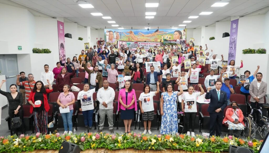 Fotografía grupal en un auditorio con decenas de asistentes de pie entre las filas de asientos. Varias personas sostienen carteles y fotografías impresas durante un acto público. En la parte frontal del escenario hay arreglos florales y al fondo un mural de gran formato.