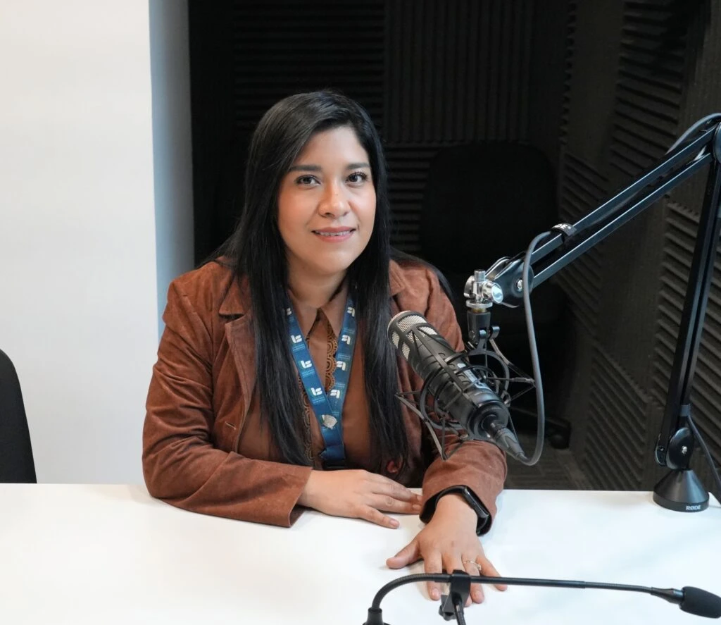 Persona sentada frente a una mesa en un estudio de grabación o cabina de pódcast, con un micrófono profesional montado en brazo articulado. La persona porta una acreditación institucional alrededor del cuello y viste una chaqueta color marrón. El espacio cuenta con paneles acústicos oscuros en las paredes y equipamiento de audio visible en primer plano.