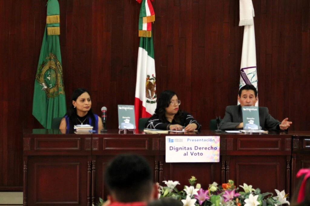 Mesa de presentación en un auditorio durante un evento académico o institucional. Dos personas participan en el panel y utilizan micrófonos de sobremesa. Sobre la mesa se exhiben ejemplares de una publicación titulada “Dignitas Derecho al Voto”, mientras al fondo se aprecia un revestimiento de madera.