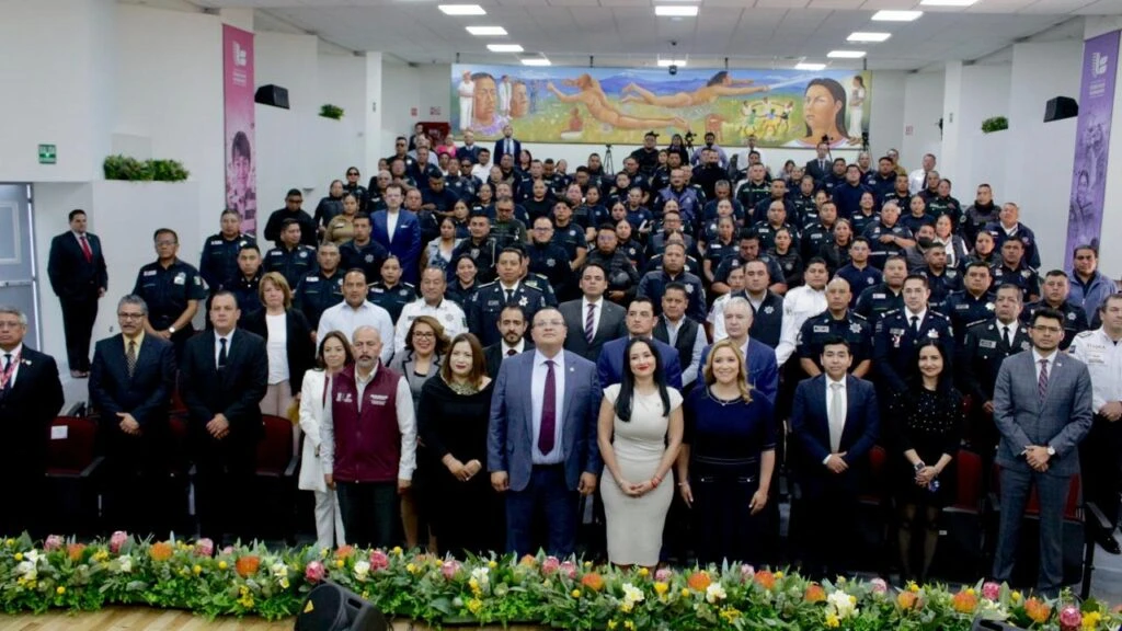 Fotografía grupal en un auditorio institucional con numerosas personas distribuidas en varias filas. En primer plano se encuentran autoridades, personal de instituciones públicas y participantes del evento; al fondo se observan integrantes de corporaciones de seguridad uniformados. El escenario está decorado con arreglos florales y un mural de gran formato en la pared posterior.