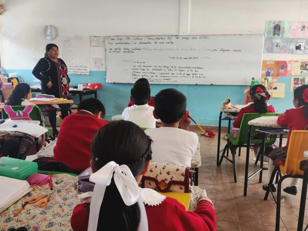 Patio central techado de una escuela con una amplia cubierta metálica arqueada. A los costados se observan edificios escolares de un nivel pintados en tonos amarillo y blanco, mientras varias personas transitan o permanecen agrupadas en diferentes áreas del plantel. En el centro se aprecia una cancha de usos múltiples con tablero de baloncesto.