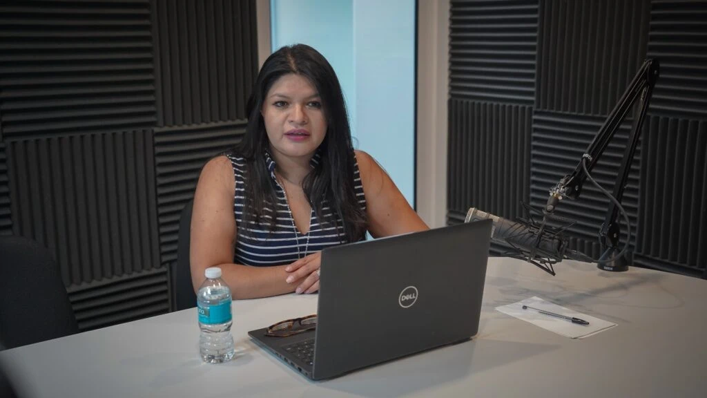 Persona sentada frente a una mesa de trabajo en un estudio de grabación equipado con paneles acústicos en las paredes. Sobre la mesa se observan un ordenador portátil, un micrófono profesional montado en brazo articulado, una botella de agua, unas gafas y una libreta con bolígrafo. La imagen muestra un entorno preparado para la grabación de un pódcast, entrevista o contenido de difusión institucional.