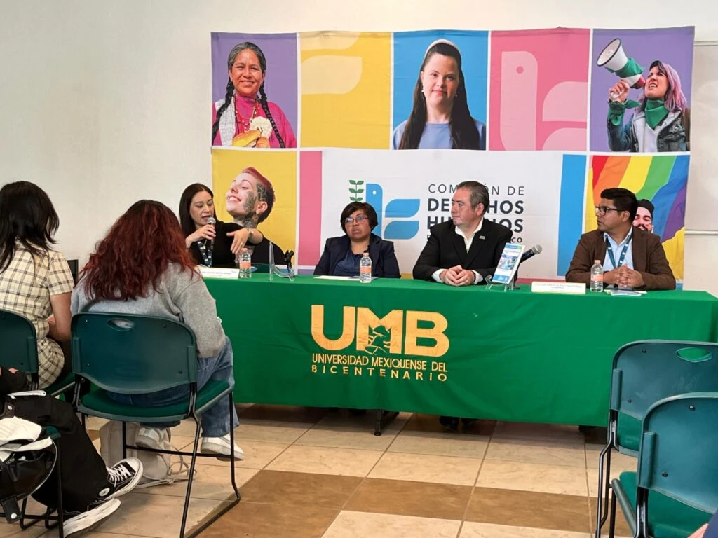 Panel de diálogo realizado en la Universidad Mexiquense del Bicentenario (UMB). Cuatro personas participan en una mesa de trabajo cubierta con un mantel verde que muestra el logotipo institucional de la universidad