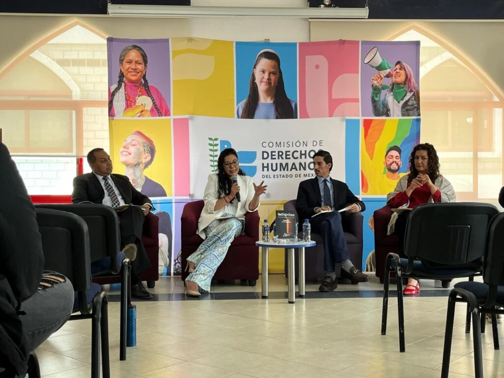 Conversatorio institucional con cuatro participantes sentados en sillones individuales. Una persona realiza una intervención mediante un micrófono mientras las demás escuchan y toman notas. En el espacio se observan materiales de consulta, botellas de agua y elementos gráficos de la Comisión de Derechos Humanos del Estado de México y de la Universidad Autónoma del Estado de México.