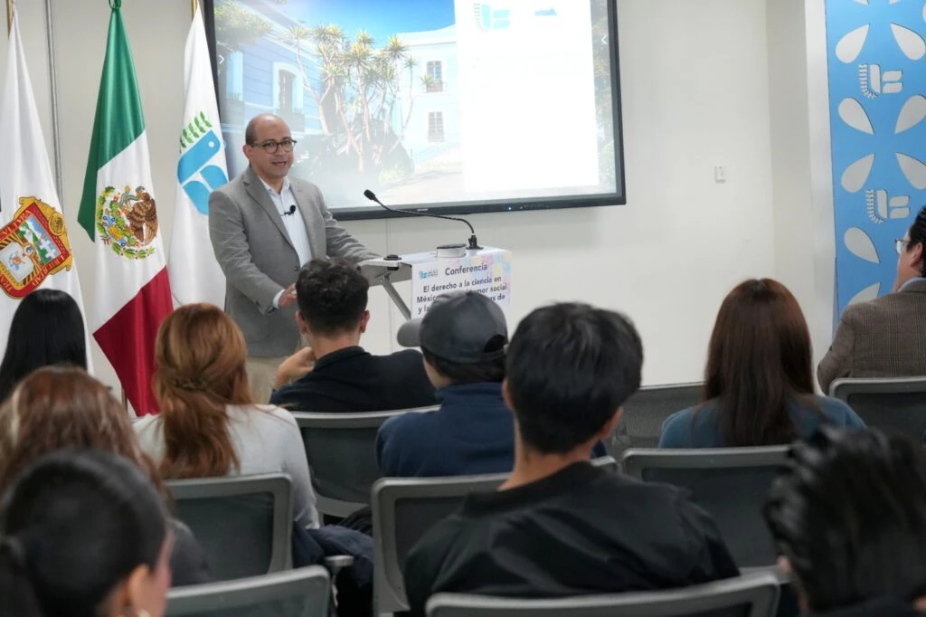 Persona expositora dirigiéndose al público durante una conferencia en una sala de reuniones, mientras las personas asistentes observan desde sus asientos junto a las banderas institucionales.