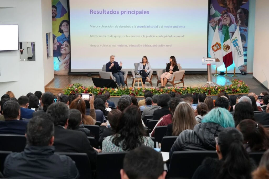 Auditorio con asistencia numerosa durante la presentación de resultados de un informe, con tres personas participando en una mesa de diálogo sobre el escenario y una proyección al fondo.