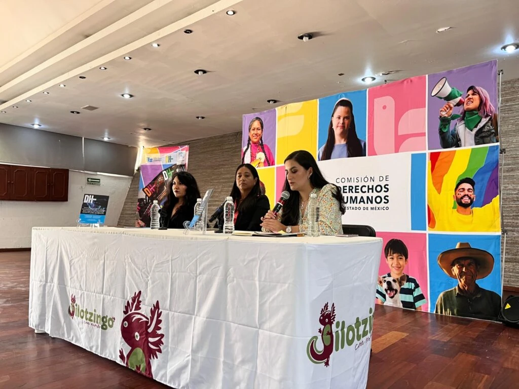 Panel informativo celebrado en un auditorio, con tres personas sentadas detrás de una mesa cubierta con un mantel institucional. Al fondo se observa un mural colorido de la Comisión de Derechos Humanos del Estado de México con fotografías ilustrativas de diversidad e inclusión.