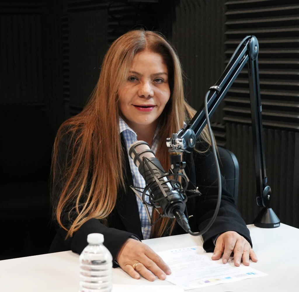 Persona sentada frente a un micrófono profesional en un estudio de grabación o radiodifusión, con paneles acústicos negros al fondo. Sobre la mesa hay documentos impresos y una botella de agua, mientras el brazo articulado del micrófono ocupa parte del primer plano de la imagen.