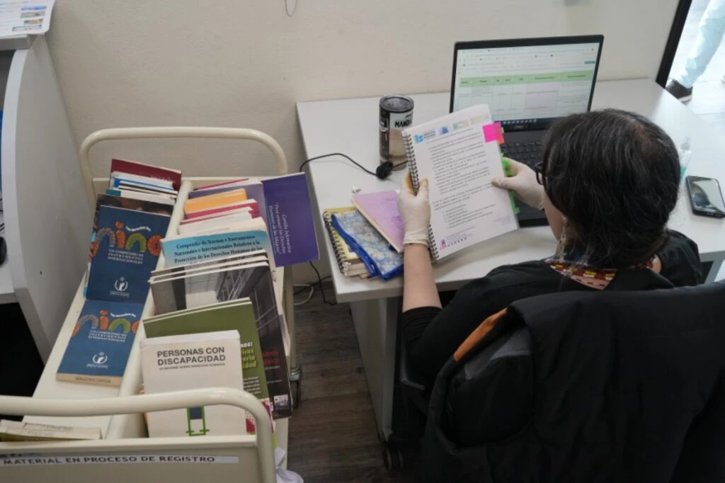 Espacio de trabajo en una biblioteca donde una persona consulta un documento impreso junto a un ordenador portátil. En primer plano se observa un carro con libros, manuales y materiales documentales en proceso de registro o revisión.