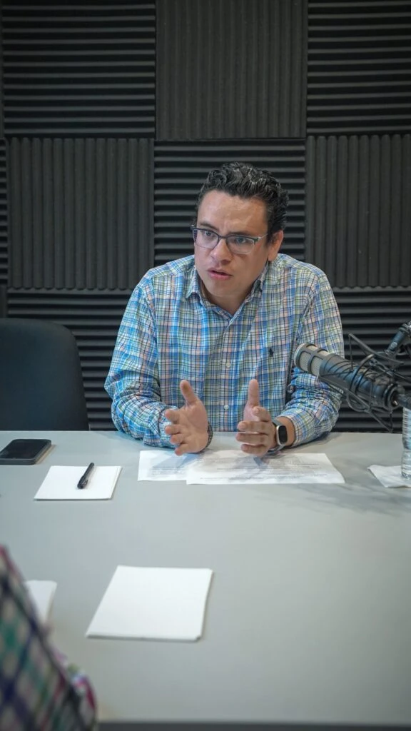 Persona sentada en una mesa de grabación o reunión, frente a un micrófono profesional montado en brazo articulado. Sobre la mesa hay documentos impresos, una libreta, un teléfono móvil y una botella de agua. El fondo está cubierto por paneles acústicos negros dispuestos en patrón geométrico, característicos de un estudio de audio o radiodifusión.