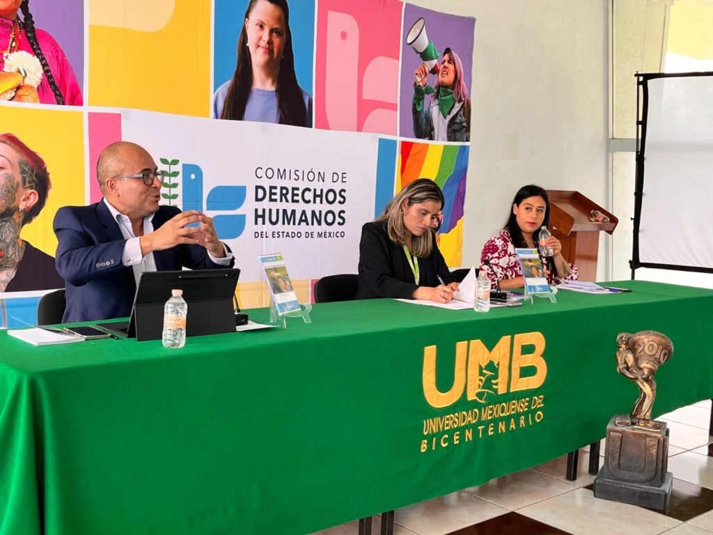 Mesa de registro y difusión con publicaciones impresas de la Comisión de Derechos Humanos del Estado de México. Varias personas consultan materiales informativos y utilizan dispositivos móviles en el acceso de un edificio iluminado por luz natural.