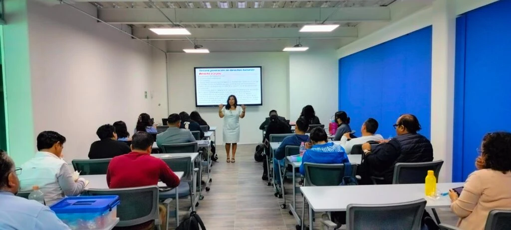 Aula de capacitación con varias personas sentadas en mesas mientras una expositora realiza una presentación frente a una pantalla de proyección. El espacio cuenta con mobiliario para formación académica y equipo audiovisual.