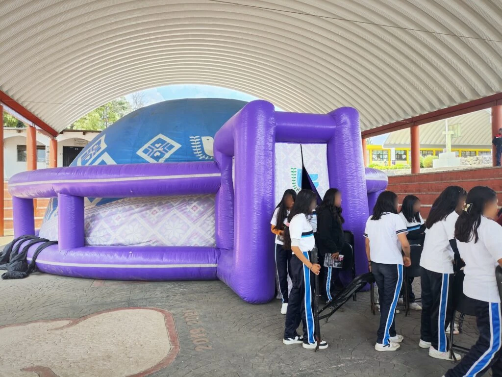 Estructura inflable de gran tamaño con forma de domo instalada en un espacio cubierto. Varias personas hacen fila junto a la entrada del inflable para participar en una actividad educativa o informativa. El domo presenta elementos gráficos institucionales en tonos azules y morados.