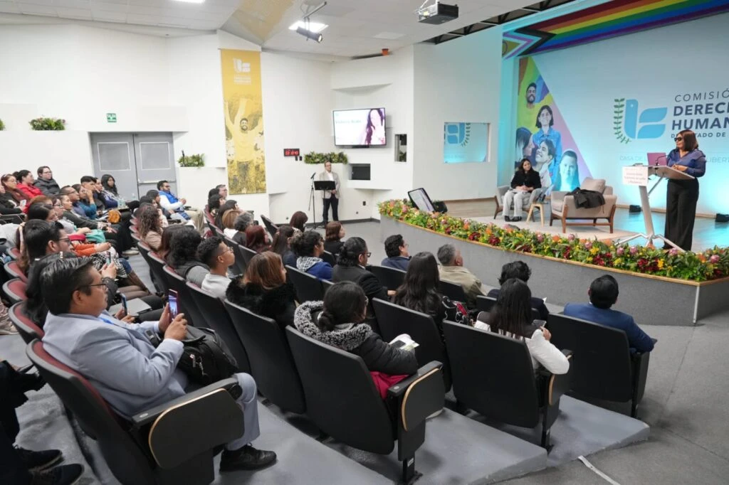 Auditorio con público asistente durante un evento organizado por la Comisión de Derechos Humanos del Estado de México. En el escenario, una persona realiza una intervención desde un atril mientras otras personas participan en una mesa de diálogo. El foro cuenta con una escenografía institucional y arreglos florales al frente.
