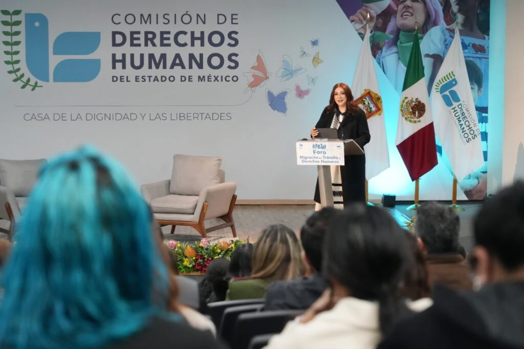 Persona realizando una ponencia desde un atril en el escenario de la Casa de la Dignidad y las Libertades. Detrás se observan las banderas de México y de la Comisión de Derechos Humanos del Estado de México, así como la identidad visual del evento y público asistente en primer plano.