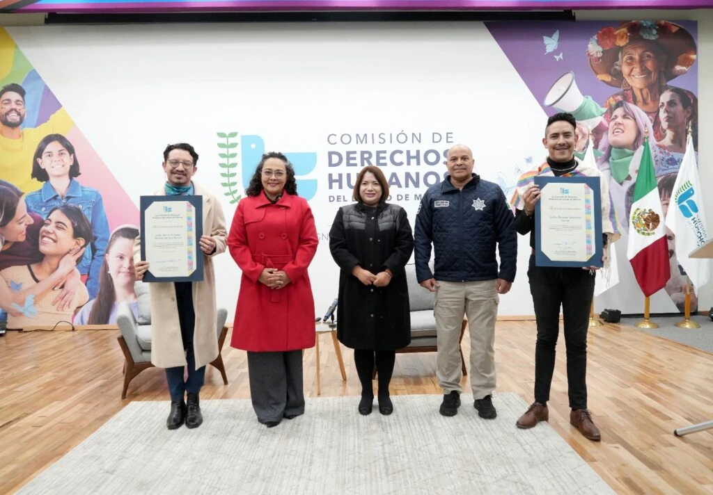Presentación pública de una guía para la atención de personas LGBTTTI+ en un auditorio. Una persona interviene desde un atril mientras dos participantes permanecen sentados en sillones sobre el escenario. En la pantalla principal se lee “Presentación de la Guía para la atención de las personas LGBTTTI+”. El público ocupa las butacas frente al escenario decorado con arreglos florales.