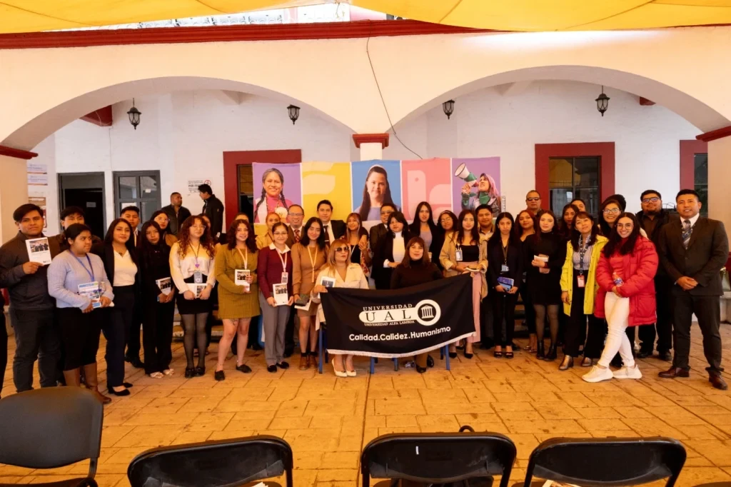 Fotografía grupal tomada en un patio cubierto durante una actividad académica o institucional. Un numeroso grupo de personas posa detrás de una mesa y frente a un panel gráfico con imágenes relacionadas con derechos humanos. En el centro del grupo se exhibe una manta de la Universidad UAEM, mientras varias personas sostienen materiales impresos y credenciales de identificación.