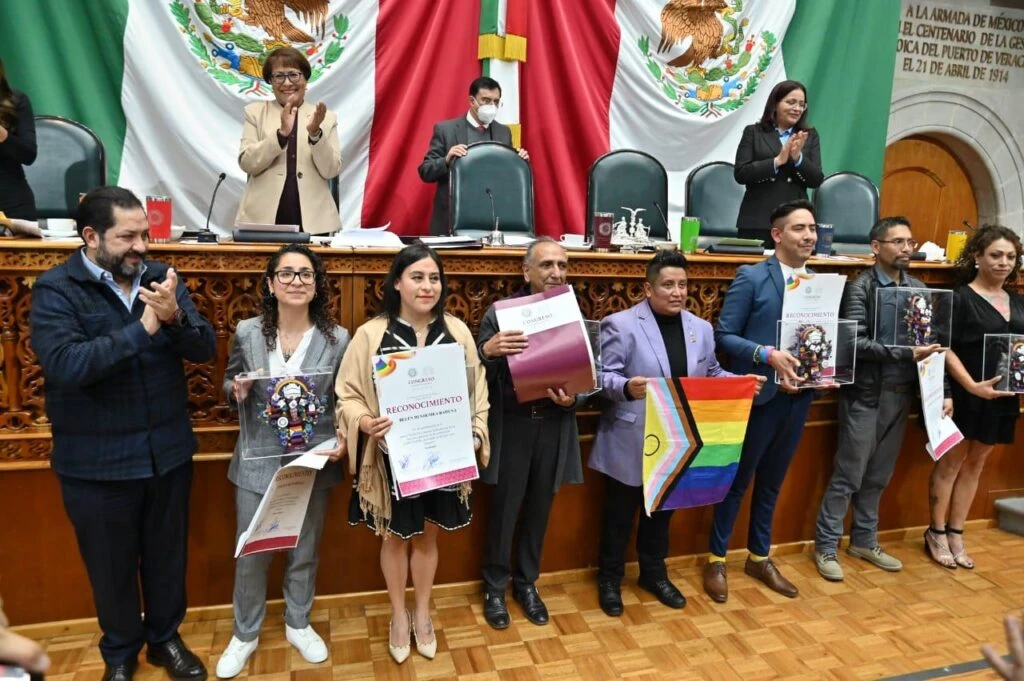 Fotografía institucional tomada durante una ceremonia de reconocimiento en un recinto parlamentario. Cuatro personas posan frente al estrado principal; dos sostienen diplomas y piezas conmemorativas de acrílico. En el fondo se distinguen las banderas nacionales y el mobiliario de la mesa directiva.