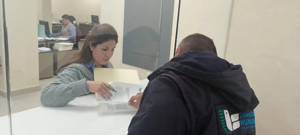 Orientación administrativa en una ventanilla de atención al público. Una trabajadora sostiene una carpeta y explica el contenido de varios documentos a una persona usuaria que revisa un expediente. El espacio corresponde a un área de trámites con mobiliario de oficina y señalización interna.