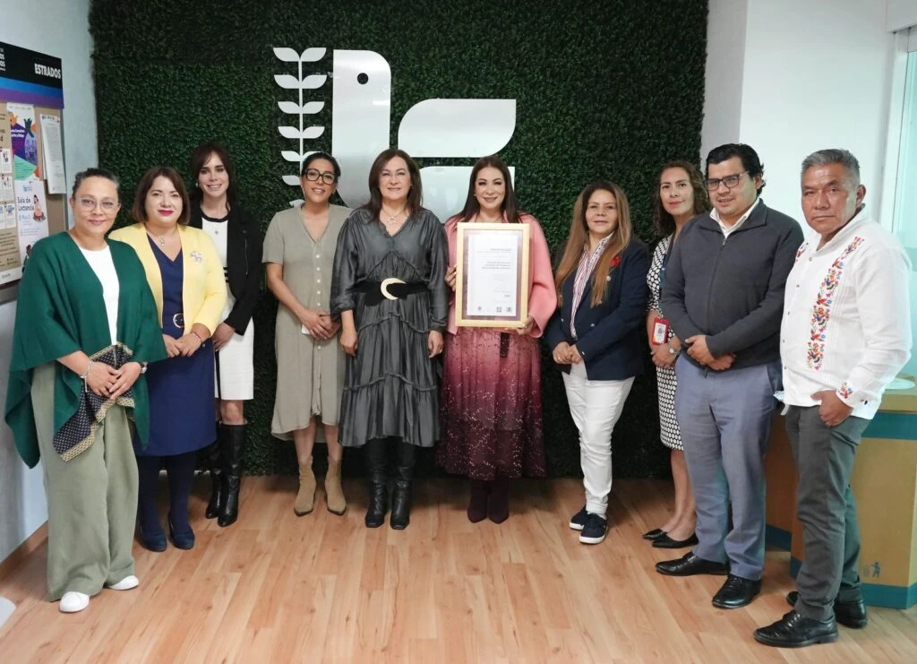 Fotografía grupal en instalaciones de la Comisión de Derechos Humanos del Estado de México. Varias personas posan frente a un muro verde con el logotipo institucional. En el centro, dos personas sostienen un reconocimiento enmarcado mientras el resto del grupo se distribuye a ambos lados para la fotografía oficial.