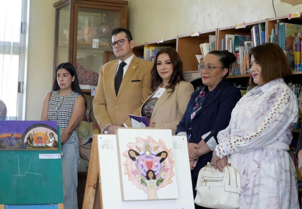 Actividad cultural y educativa realizada en una biblioteca escolar. Varias personas se encuentran de pie junto a estanterías con libros y materiales didácticos. En primer plano se observan ilustraciones artísticas expuestas sobre caballetes, mientras las personas asistentes participan en una presentación relacionada con derechos humanos y expresión artística.