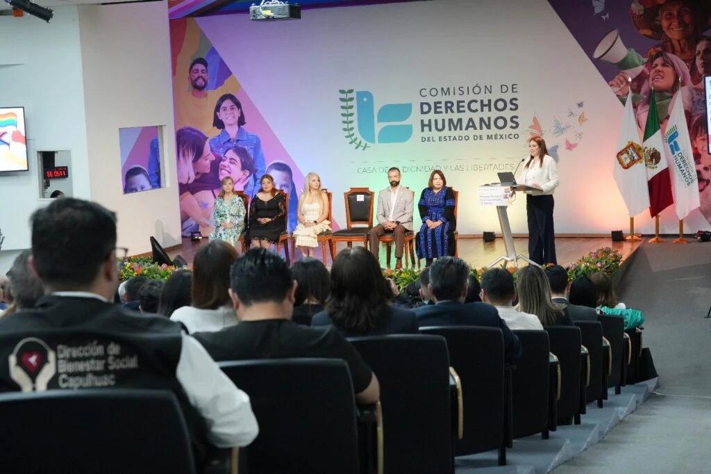 Auditorio de la Casa de la Dignidad y las Libertades durante una jornada conmemorativa relacionada con los derechos humanos de las personas LGBTIQ+. En el escenario, varias personas participan en una mesa de honor mientras una ponente interviene desde un atril. Al frente del escenario hay arreglos florales y, al fondo, se observan los logotipos de la Comisión de Derechos Humanos del Estado de México, así como las banderas institucionales. El público ocupa la mayor parte de las butacas del recinto.