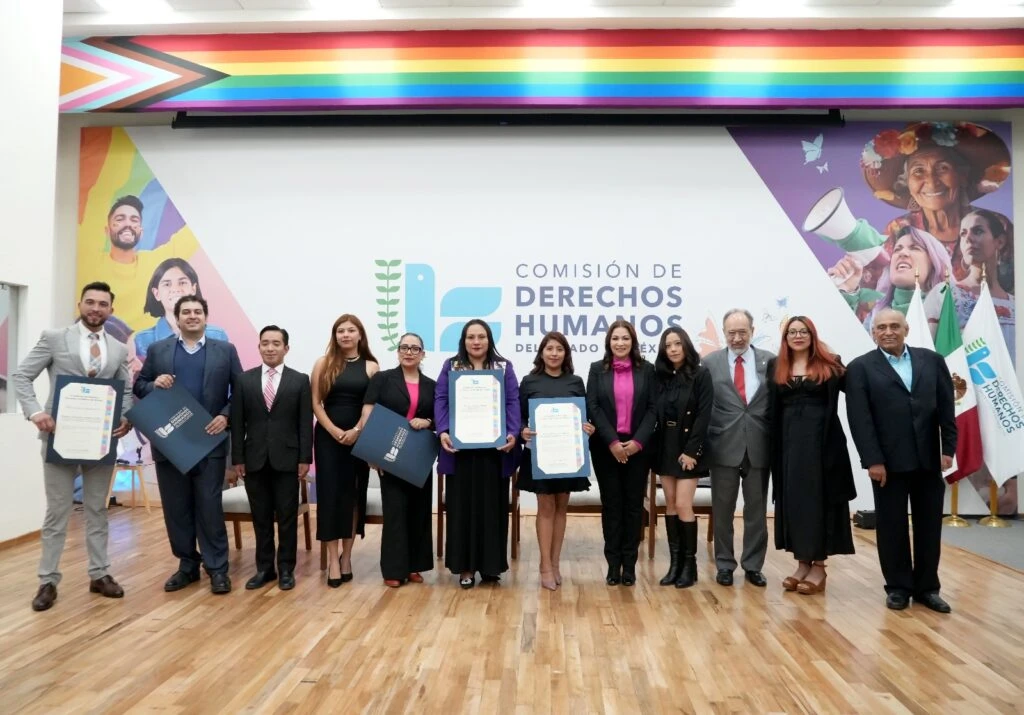 Fotografía oficial de un grupo de participantes y autoridades sobre el escenario de un auditorio. Varias personas posan frente al logotipo de la Comisión de Derechos Humanos del Estado de México; algunas sostienen reconocimientos y carpetas institucionales. Al fondo se observan las banderas de México y de la institución, así como elementos gráficos relacionados con la diversidad e inclusión.
