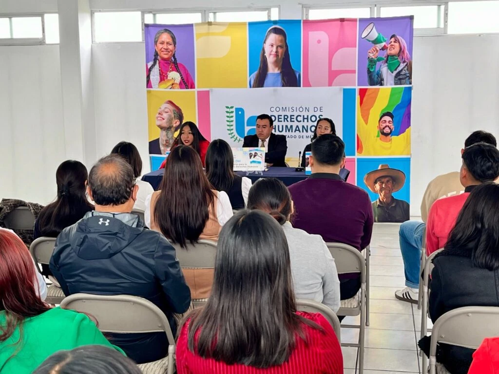 Vista general de una conferencia o presentación académica. Personas asistentes permanecen sentadas en filas de sillas observando a las y los ponentes ubicados al frente de la sala. En el fondo se encuentra un panel institucional de la Comisión de Derechos Humanos del Estado de México con imágenes alusivas a la diversidad social y cultural.