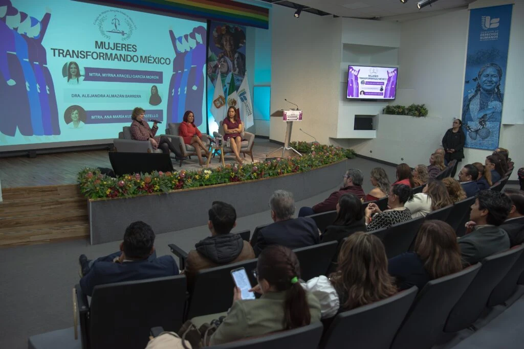 Vista general de un auditorio durante el conversatorio “Mujeres Transformando México”. En el escenario hay tres participantes sentadas junto a un atril, mientras una gran pantalla muestra el título del evento. El público ocupa las butacas observando la actividad, y el escenario está decorado con flores y elementos institucionales.