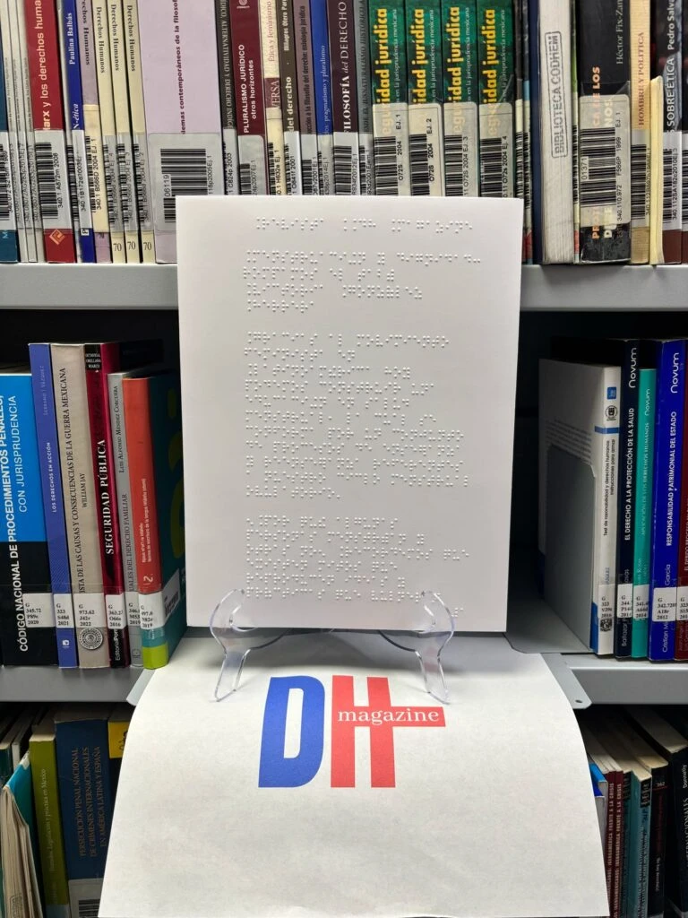 Publicación en sistema braille colocada sobre un soporte transparente dentro de una biblioteca. Al fondo se observan estanterías con libros especializados catalogados mediante códigos de barras. En la parte inferior aparece una carpeta o publicación con el logotipo “DH magazine”, relacionada con derechos humanos y accesibilidad.