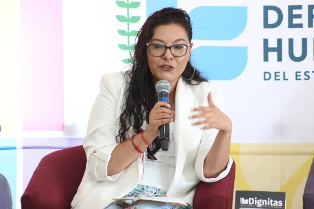 Persona participante durante una intervención en un foro o conversatorio sobre derechos humanos. Se encuentra sentada en un sillón color burdeos, sosteniendo un micrófono mientras realiza una exposición. Al fondo aparecen elementos gráficos institucionales, incluido un logotipo relacionado con derechos humanos y materiales de comunicación del evento.