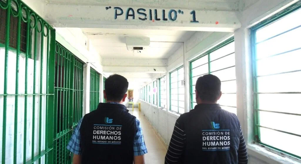 Pasillo interior de un centro de reclusión identificado con el rótulo “PASILLO 1” en la parte superior. A ambos lados se observan estructuras metálicas pintadas de verde y ventanas que permiten la entrada de luz natural