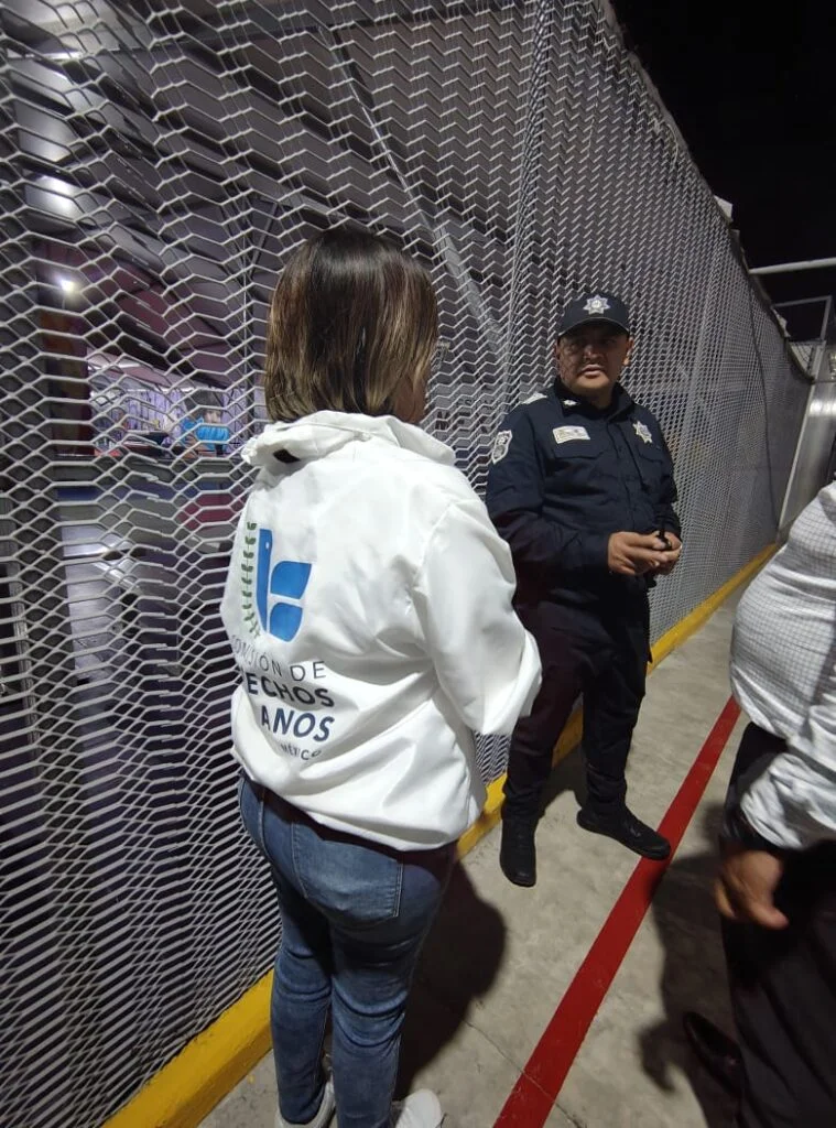 Personal de la Comisión de Derechos Humanos del Estado de México dialoga con un elemento de seguridad en una zona de circulación delimitada por una malla metálica. La escena se desarrolla en un espacio interior con iluminación artificial, como parte de labores de observación y supervisión institucional.
