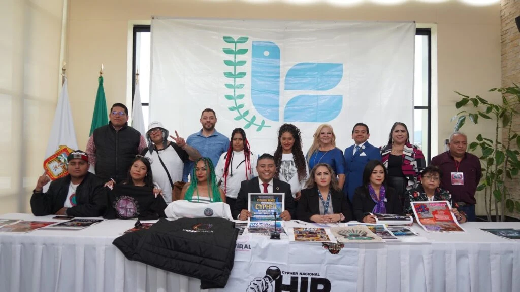 Fotografía grupal de participantes de un evento realizado en un salón de conferencias. Las personas posan detrás de una mesa con publicaciones, prendas promocionales y materiales informativos, frente a un fondo institucional.