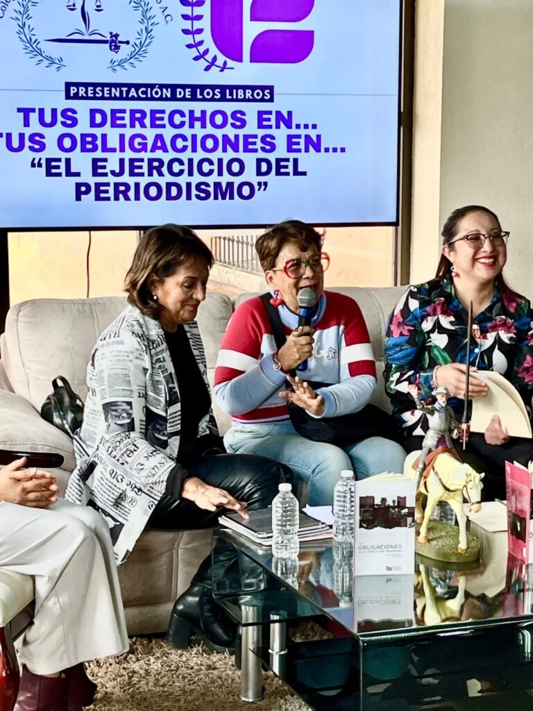 Acercamiento a una mesa de conversación durante la presentación de libros. Una persona interviene con micrófono mientras otras participantes escuchan y consultan materiales impresos sobre la mesa de cristal.