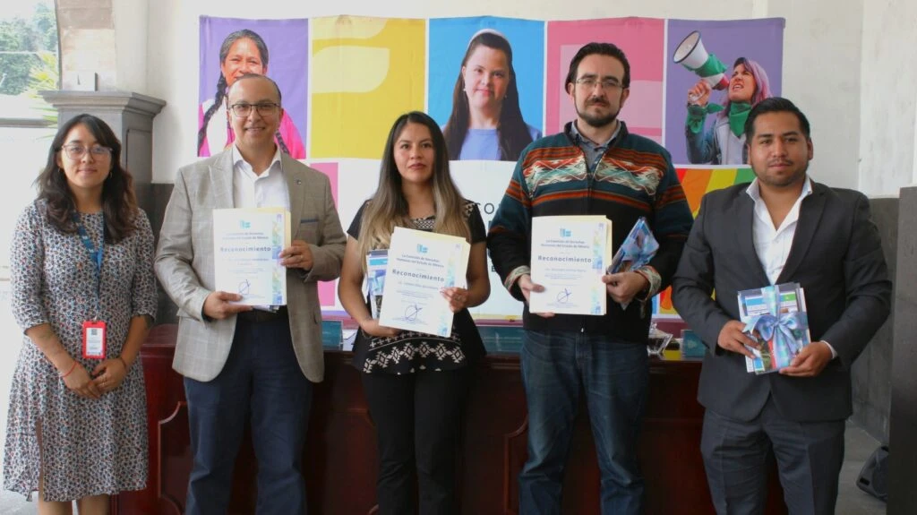 Texto alternativo: Grupo de cinco personas de pie frente a una mesa y una lona institucional de la Comisión de Derechos Humanos del Estado de México. Varias personas sostienen reconocimientos impresos y materiales gráficos, con un fondo de paneles coloridos y fotografías.