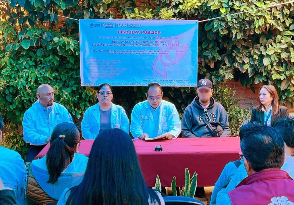 Texto alternativo: Evento de disculpa pública desarrollado al aire libre ante un grupo de asistentes. Varias personas están sentadas frente al público detrás de una mesa rectangular. En la parte posterior se aprecia una lona informativa con el título “Disculpa Pública”, rodeada de vegetación y un muro de ladrillo.