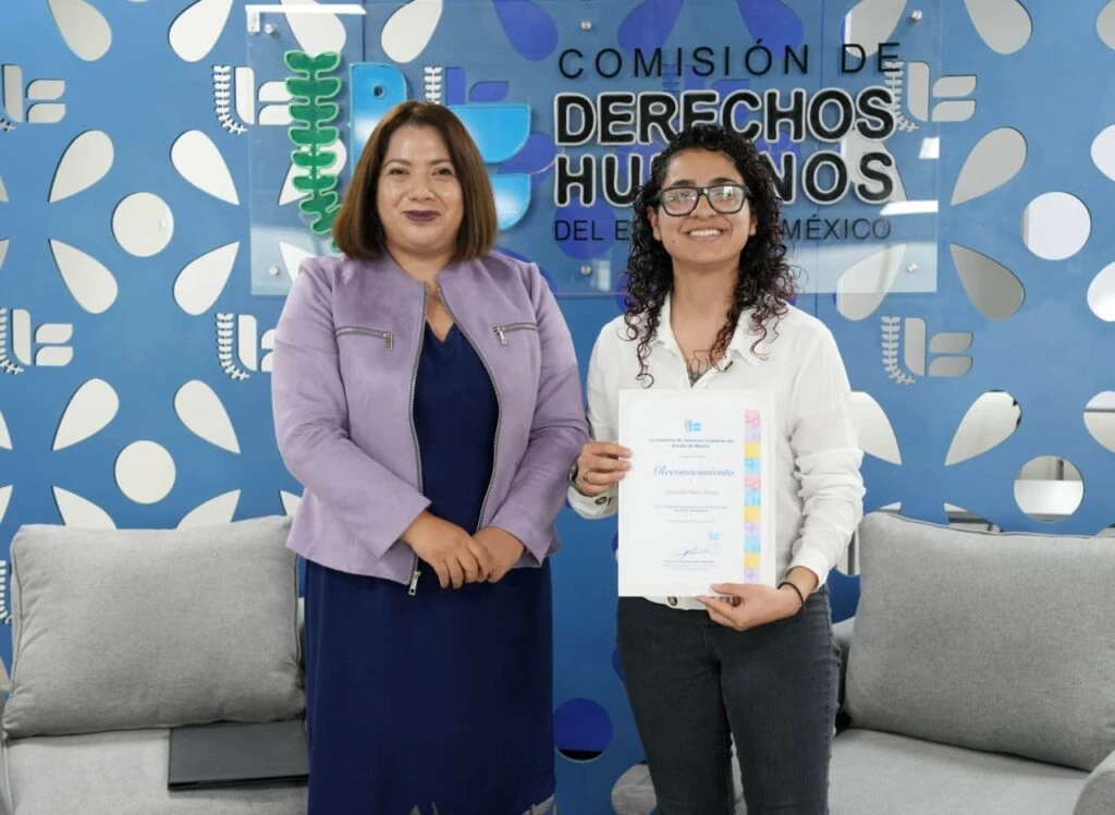 Texto alternativo: Dos personas posan en una sala institucional frente a un muro azul con el texto “Comisión de Derechos Humanos del Estado de México”. Una de las personas sostiene un reconocimiento impreso; alrededor se observan sillones grises y elementos decorativos con logotipos institucionales.