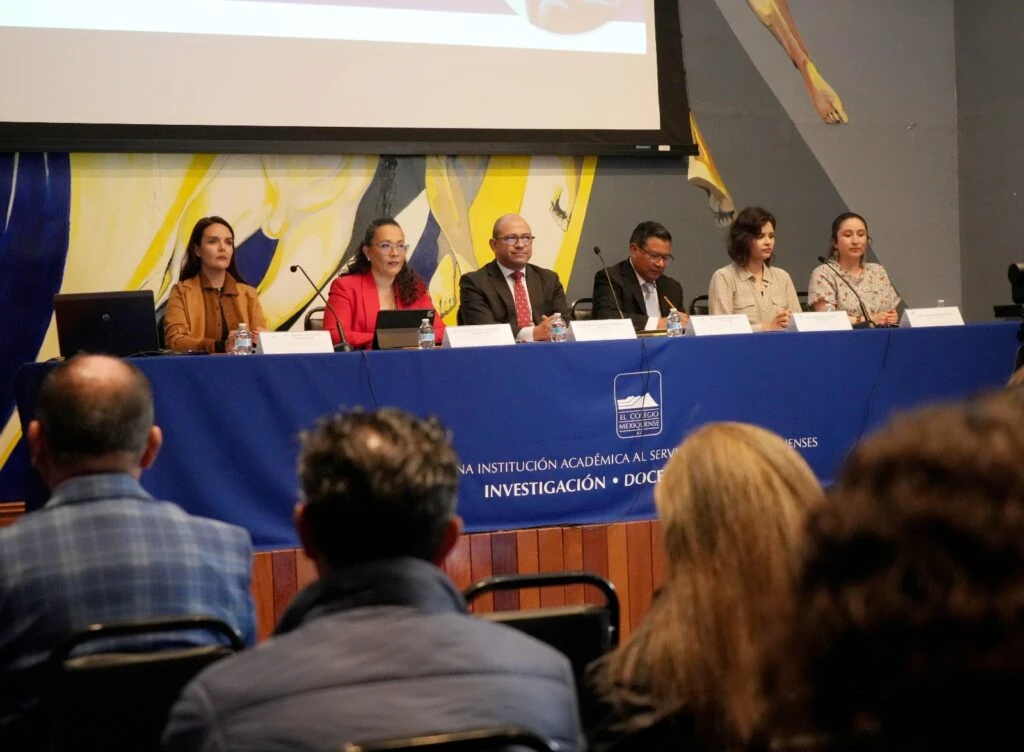 Texto alternativo: Mesa de panel en un auditorio con seis personas sentadas detrás de una mesa cubierta con mantel azul. Sobre la mesa hay micrófonos, botellas de agua, identificadores y equipos electrónicos. Al fondo se aprecia una pantalla de proyección y parte de un mural. En el mantel se observa el logotipo de “El Colegio Mexiquense” y texto relacionado con investigación y docencia.