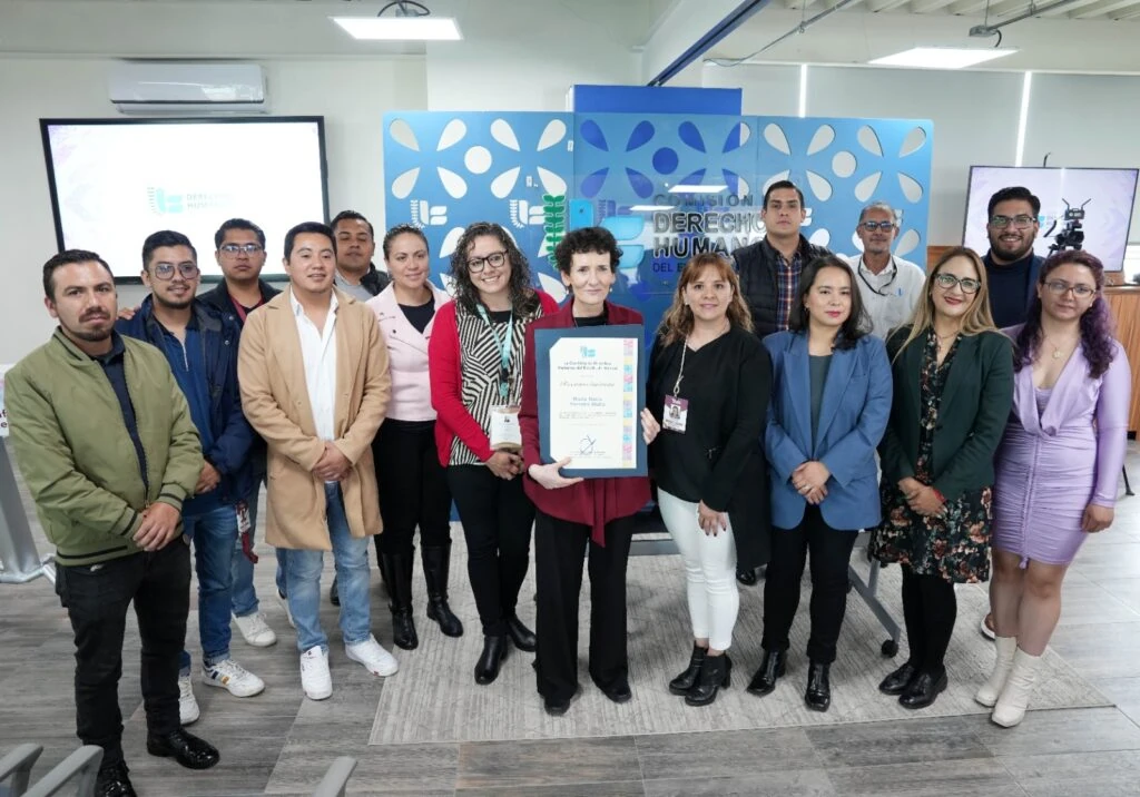 Texto alternativo: Grupo de personas de pie en una sala institucional frente a un panel azul con el texto “Comisión de Derechos Humanos del Estado de México”. En el centro, una persona sostiene un reconocimiento enmarcado mientras el resto del grupo posa alrededor. Al fondo se observan pantallas, mobiliario y equipo audiovisual.