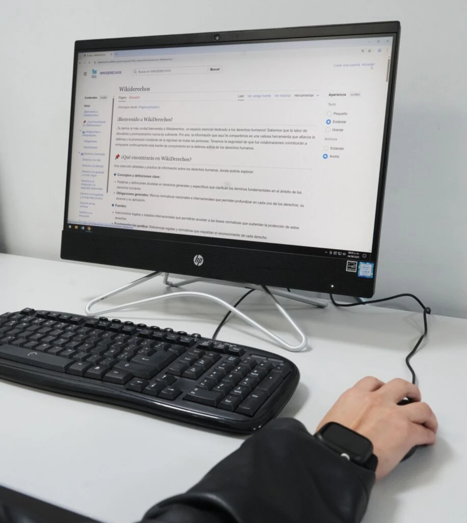 Monitor HP colocado sobre un escritorio blanco junto a un teclado y un ratón. En la pantalla se visualiza el sitio “WikiDerechos”, una plataforma digital relacionada con derechos humanos, con un mensaje de bienvenida y un menú de navegación lateral. En primer plano se observa una mano utilizando el ratón y una pulsera o dispositivo electrónico en la muñeca. El entorno corresponde a un espacio de trabajo o centro de cómputo con paredes blancas y mobiliario de oficina.