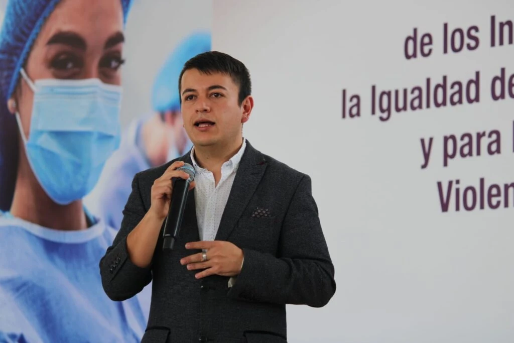Persona sosteniendo un micrófono durante una conferencia o presentación en un espacio interior. Al fondo se observa una pantalla o muro con texto parcialmente visible relacionado con la igualdad y la prevención de la violencia.