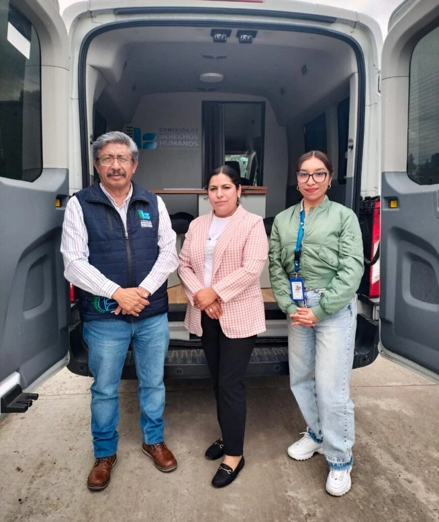 Tres personas posan frente a una unidad móvil de la Comisión de Derechos Humanos del Estado de México con las puertas traseras abiertas. En el interior de la unidad se observa un módulo de atención y el logotipo institucional. La fotografía fue tomada en un espacio exterior pavimentado.