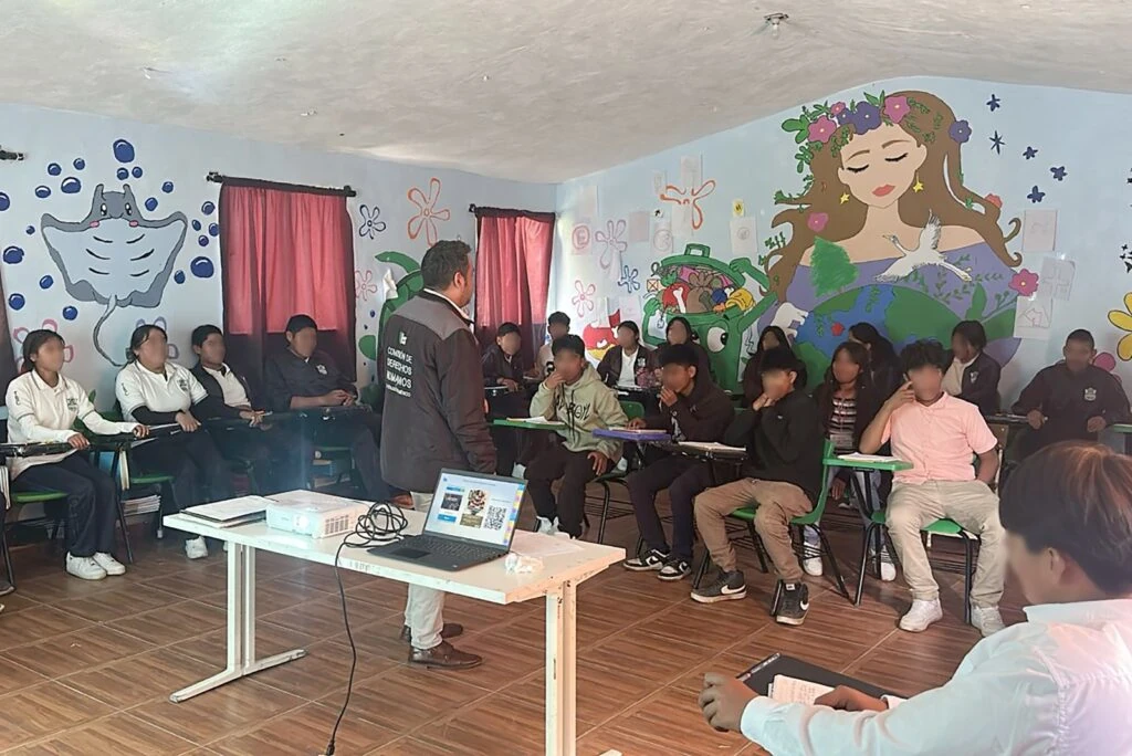Grupo de estudiantes sentados en pupitres dentro de un aula con murales infantiles en las paredes. Al frente, una persona con chaleco institucional de la Comisión de Derechos Humanos del Estado de México realiza una exposición apoyada por una mesa con proyector y computadora portátil