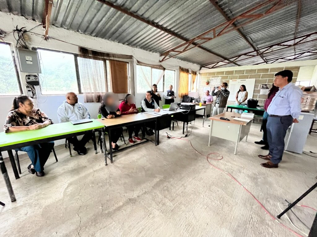 Reunión o sesión informativa en una sala amplia con techo de lámina y varias mesas acomodadas en forma de trabajo. Personas sentadas observan a una persona de pie que expone frente al grupo.