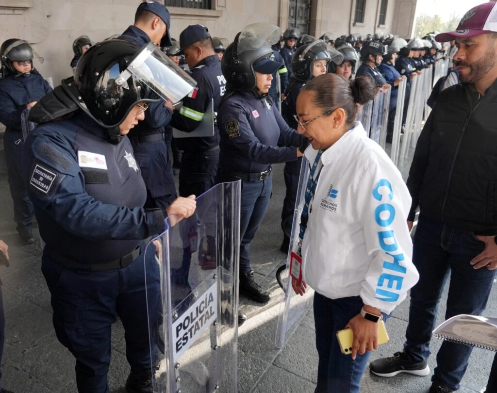 Personal de policía estatal sostiene escudos antimotines en un corredor exterior, mientras una persona con chaqueta blanca institucional revisa el equipo durante una supervisión operativa.