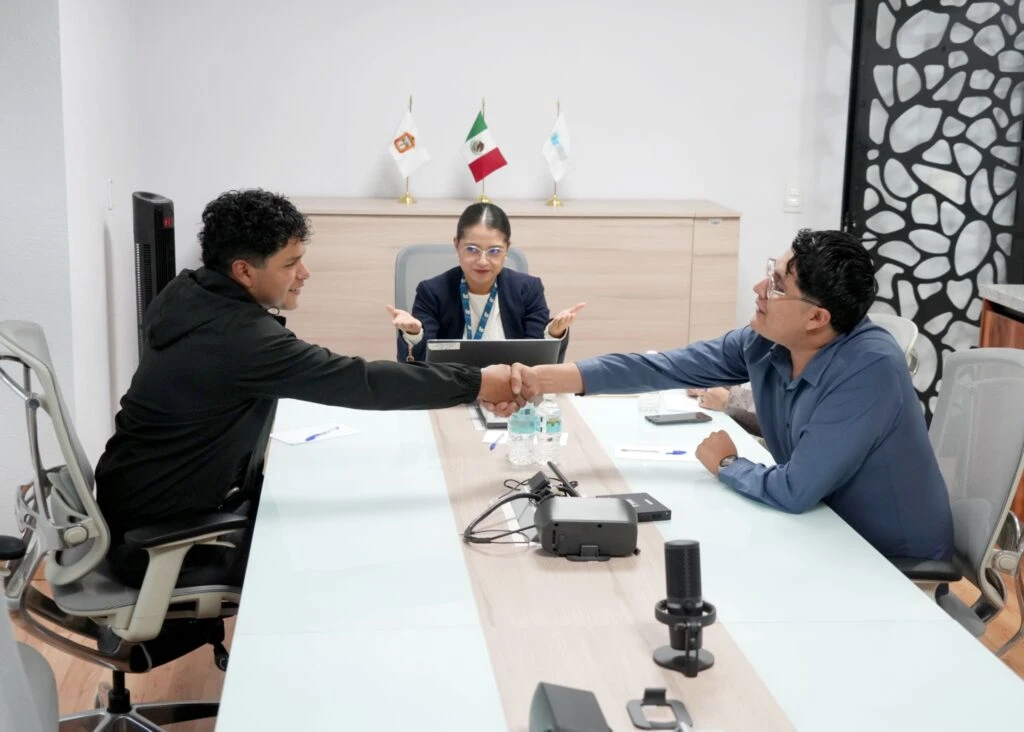 Cuatro personas participan en una reunión alrededor de una mesa de juntas en una oficina. Una persona ubicada al centro realiza una intervención mientras utiliza un ordenador portátil; sobre la mesa hay botellas de agua, material de oficina y equipo para videoconferencia. Al fondo se observan banderas institucionales y una mampara decorativa.